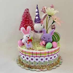 May include: An Easter-themed decorative centerpiece featuring a white ceramic bunny wearing a purple polka-dot hat, surrounded by other bunnies, chicks, eggs, and flowers. The base is a layered cake design with patterned ribbons and gold trim.