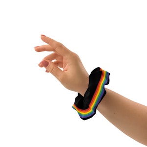 May include: A black scrunchie with a rainbow stripe design. The scrunchie is worn on a person's wrist.