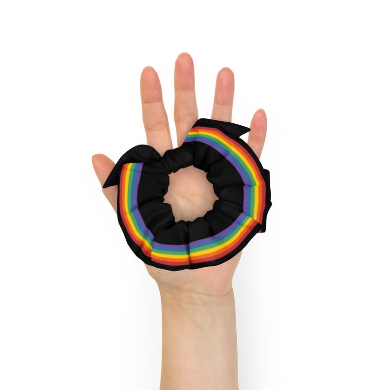 May include: A black hair scrunchie with a rainbow stripe design. The scrunchie is held in a hand.