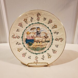 Könnte beinhalten: Ein weißer Keramikteller mit goldfarbenem Rand zeigt eine farbenfrohe Illustration von zwei Kindern mit einer Ziege und einem Wagen. Der Teller ist mit einem grünen Rand und einem Ring aus Blumen verziert. Der Text auf dem Teller lautet "Little Greenways Almanac 1984, Capricornus, The Goat".