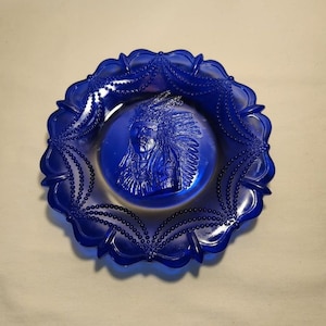 May include: Cobalt blue glass plate with a raised design of a Native American man in a headdress. The plate has a scalloped edge with a beaded design.