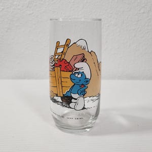 Puede incluir: Un vaso de vidrio transparente con una imagen de dibujos animados de un Pitufo empujando un carro lleno de materiales de construcción. El Pitufo lleva un sombrero azul y pantalones azules. El texto en la parte inferior del vaso dice "SEPP. DM. INTL."