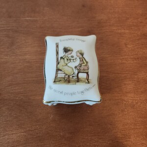 May include: White porcelain trinket box with a gold trim. The box features a picture of two girls sitting on chairs, holding cups. The text on the box reads "Friendship brings the nicest people together."
