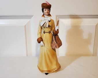 May include: A porcelain figurine of a woman wearing a yellow coat with brown fur trim, a white bow, and a brown hat with a pink feather. She is holding a brown handbag with a floral pattern.