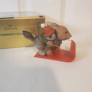 May include: A Hallmark Trimmer Collection ornament featuring a brown rabbit pulling a red sled with a smaller brown rabbit wearing a green sweater and red hat.