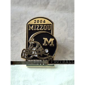 May include: A gold and black metal lapel pin featuring a football helmet with the letter M. The pin displays the text 2004 and MIZZOU at the top, with the word FOOTBALL partially visible at the bottom. The design includes a black background.