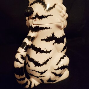 May include: A ceramic tiger-shaped container with a lid, featuring a white base and black stripes. The tiger is in a seated position with its tail curled. The container has a black nose and ears. The overall design is whimsical and decorative.