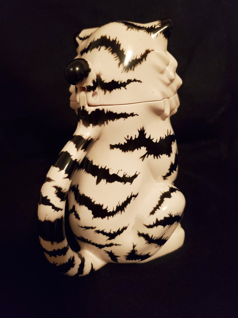 May include: A ceramic tiger-shaped container with a lid, featuring a white base and black stripes. The tiger is in a seated position with its tail curled. The container has a black nose and ears. The overall design is whimsical and decorative.