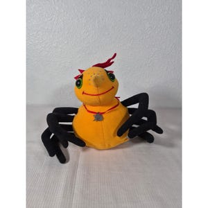Puede incluir: Una araña de peluche con cuerpo amarillo, patas negras y detalles rojos. La araña tiene ojos verdes grandes, boca sonriente y un collar rojo con un colgante plateado. El juguete está diseñado para ser amigable y acogedor.