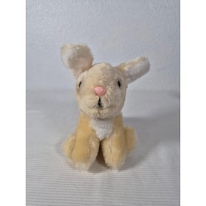 Puede incluir: Un peluche de conejo en posición sentada. El conejo tiene un cuerpo de color beige claro, detalles blancos y una nariz rosa. Sus orejas están erguidas y sus ojos son negros.