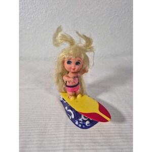 Peut inclure: Une petite poupée blonde aux yeux bleus, vêtue d'un bikini rose, se tient debout sur une planche de surf jaune, rouge et bleue. La poupée sourit et lève le bras. La planche de surf a un motif de vagues.