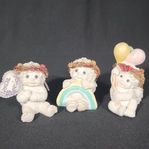 May include: Three small, white cherub figurines. One holds a heart-shaped sign that reads "You're Special." Another sits on a rainbow, and the third holds balloons. Each has a wreath and blue eyes.