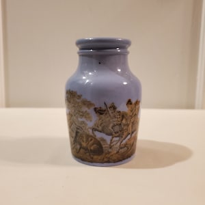 May include: A small, light blue ceramic jar with a brown and gold design of people riding horses. The jar has a simple, rounded shape and a smooth, glazed finish.