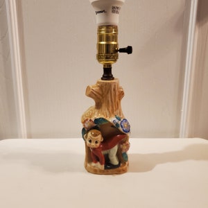 May include: A ceramic table lamp shaped like a tree trunk with a small monkey figurine peeking out from behind the trunk. The lamp has a white lightbulb and a black switch.