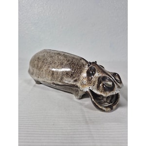 May include: A ceramic hippo figurine in shades of brown and white. The hippo is lying down with its mouth open. The figurine has a glossy finish and is likely a decorative item.