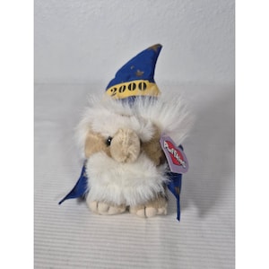 Peut inclure: Un jouet en peluche représentant un magicien avec un chapeau et une cape bleus, avec l'année "2000" sur le chapeau. Le jouet a une barbe blanche et duveteuse et un corps marron et blanc. Une étiquette "Puffkins" est attachée.