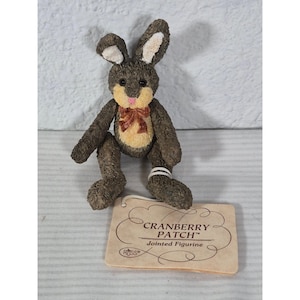 May include: A jointed figurine of a bunny rabbit with brown fur, tan belly, and pink bow. The bunny has long ears with white insides and is holding a tag that reads "Cranberry Patch Jointed Figurine".