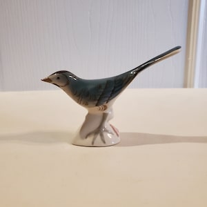 Puede incluir: Una pequeña figurita de pájaro de porcelana azul y blanca con una cola larga. El pájaro está posado sobre una pequeña base y tiene un diseño detallado.