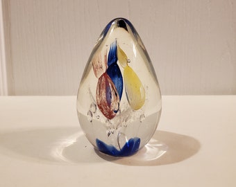May include: A clear glass paperweight with blue, yellow, and red swirls inside. The paperweight is round and has a smooth surface.