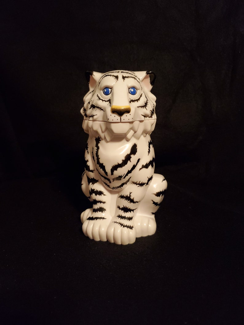 May include: A white tiger figurine with black stripes, blue eyes, and a gold nose. The ceramic tiger is sitting upright with its paws in front. The figurine has a lid on its head and is set against a black background.