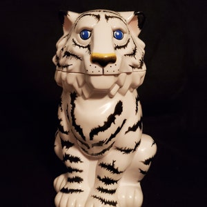 May include: A white ceramic tiger figurine with black stripes and blue eyes. The tiger has a gold nose and black accents on its ears. The figurine is sitting upright, with its paws visible. The background is black.