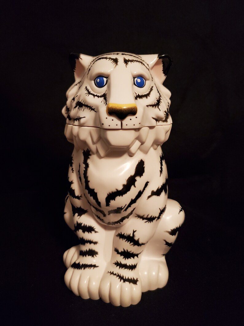 May include: A white ceramic tiger figurine with black stripes and blue eyes. The tiger has a gold nose and black accents on its ears. The figurine is sitting upright, with its paws visible. The background is black.