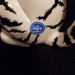 May include: A white ceramic object with black, jagged markings. A blue sticker with the text "THE GREATEST SHOW ON EARTH" is affixed to the surface. The object is set against a dark background.