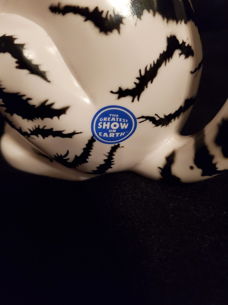 May include: A white ceramic object with black, jagged markings. A blue sticker with the text "THE GREATEST SHOW ON EARTH" is affixed to the surface. The object is set against a dark background.