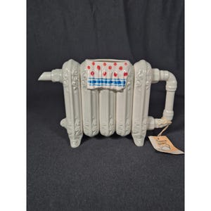 May include: A white ceramic teapot shaped like a radiator, featuring a handle and spout. The top has a decorative design with red polka dots and blue stripes. A small tag hangs from the handle.