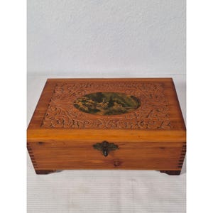 Può includere: Un portagioie in legno di forma rettangolare con coperchio incernierato. Il coperchio presenta un motivo floreale intagliato attorno a un'immagine ovale di un paesaggio con una chiesa. La scatola ha una chiusura in ottone ed è realizzata in legno marrone chiaro.