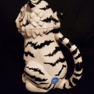May include: A white ceramic tiger-shaped pitcher with black stripes and a curved handle. The pitcher has a black and white striped tail and a small blue sticker that reads "GREATEST SHOW EARTH".