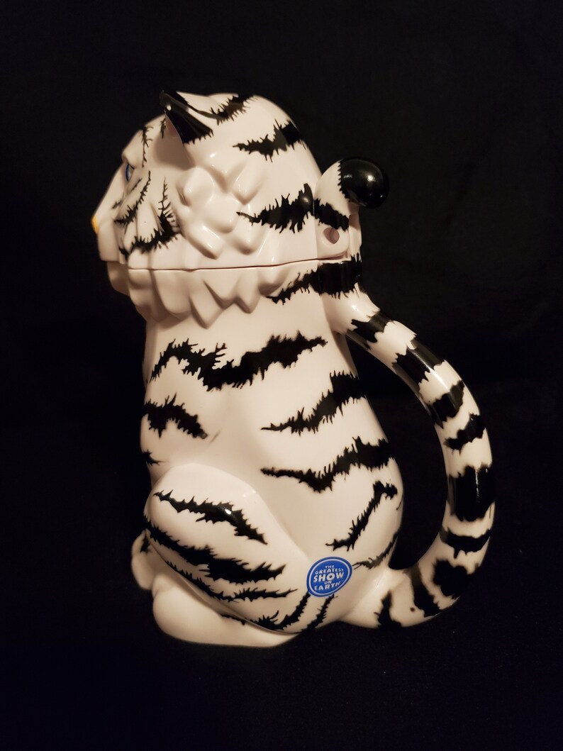 May include: A white ceramic tiger-shaped pitcher with black stripes and a curved handle. The pitcher has a black and white striped tail and a small blue sticker that reads "GREATEST SHOW EARTH".