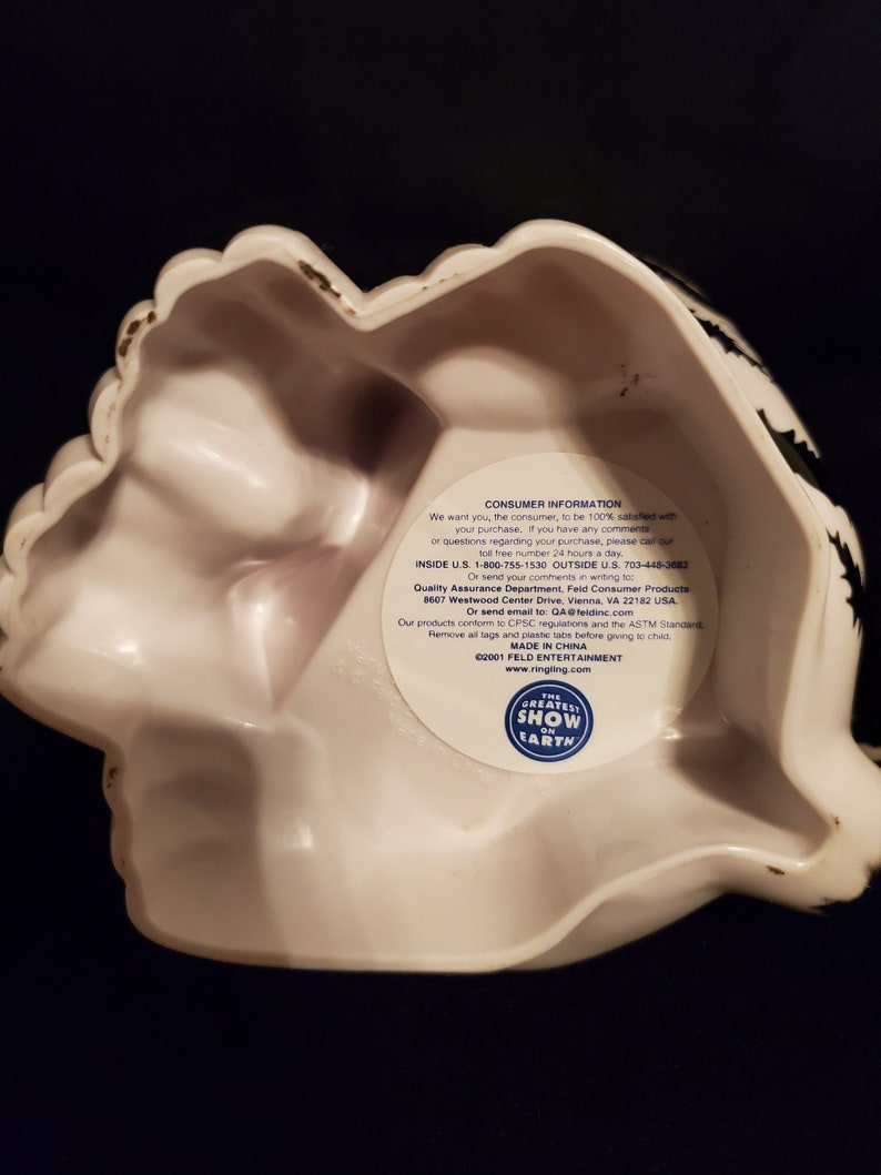 May include: White ceramic decorative bowl with a scalloped edge and black accents. The bowl has a unique shape and a sticker with text that reads "THE GREATEST SHOW ON EARTH".