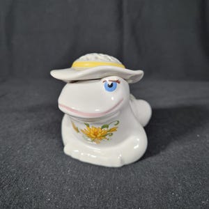 Puede incluir: Figura de rana de cerámica blanca con un sombrero blanco con una banda amarilla. La rana tiene ojos azules, boca rosa y un diseño floral amarillo y verde en el pecho. La figura está sobre una superficie gris oscuro.