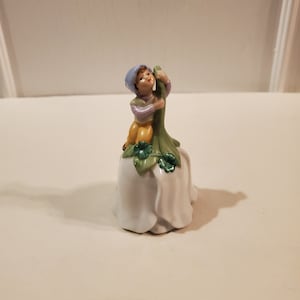 May include: A porcelain bell with a white base and a green stem. A small boy figurine sits on top of the stem, holding a green leaf. The boy is wearing a blue hat, a purple shirt, and yellow pants.