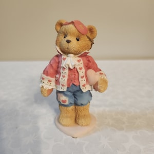 May include: A small, brown teddy bear figurine wearing a pink and white heart-patterned jacket, blue trousers, and a pink eye mask. The bear is holding a pink heart in its right paw and is standing on a pink base.