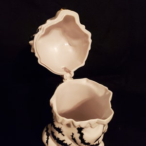 May include: A white ceramic tiger-shaped cookie jar with black stripes and a hinged lid. The lid is open, revealing the interior. The jar is set against a dark background.