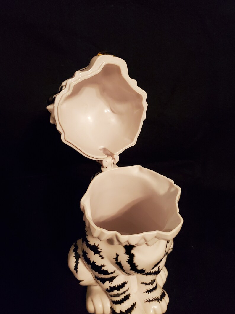 May include: A white ceramic tiger-shaped cookie jar with black stripes and a hinged lid. The lid is open, revealing the interior. The jar is set against a dark background.