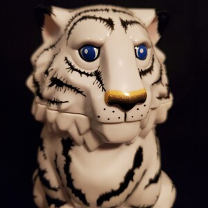 May include: A white tiger figurine with black stripes and blue eyes. The tiger has a gold-colored nose and black markings around the eyes and mouth. The figurine is set against a dark background.