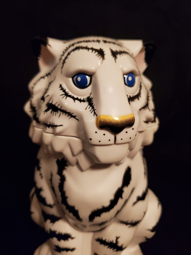 May include: A white tiger figurine with black stripes and blue eyes. The tiger has a gold-colored nose and black markings around the eyes and mouth. The figurine is set against a dark background.