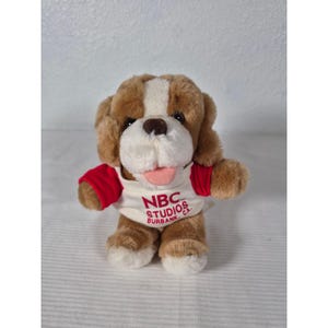 May include: A plush dog toy with brown and white fur, wearing a red-sleeved shirt. The shirt has the text "NBC STUDIOS BURBANK, CA" printed on it. The dog has a pink tongue sticking out.