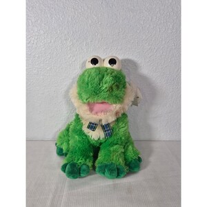 May include: A plush green frog toy with large, round eyes and a pink mouth. The frog has a cream-colored chest and a blue and green plaid bow tie. The toy is sitting upright, with its arms and legs extended.