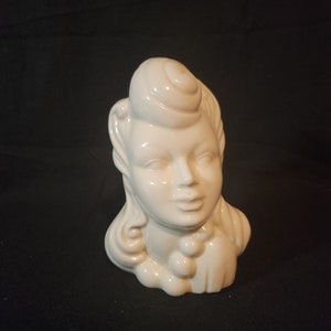 Könnte beinhalten: Weiße Keramikskulptur eines Frauenkopfes mit einem Dutt. Die Skulptur ist ein Büste und zeigt ein detailliertes Gesicht mit einer einfachen Halskette.