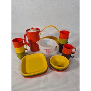 Puede incluir: Colección de utensilios de cocina vintage en varios colores. Incluye una jarra roja, un vaso medidor, platos, cuencos y tazas. Los platos y cuencos están apilados, y las tazas están en juegos. Los artículos están dispuestos sobre una superficie blanca.