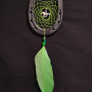 May include: A horseshoe dreamcatcher with a green web and a silver horse charm. The dreamcatcher features a green feather and green beads. The horseshoe is dark grey.