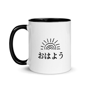 May include: White ceramic mug with a black rim and handle. The mug features a black and white illustration of a sun and the Japanese text "おはよう" which means "good morning".