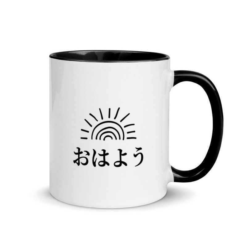 May include: White ceramic mug with a black rim and handle. The mug features a black and white illustration of a sun with a rainbow inside and the Japanese text "おはよ" which translates to "good morning".
