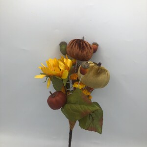 May include: A fall-themed decorative stem featuring a bright yellow sunflower, small pumpkins in shades of orange and tan, and green leaves. The stem also includes acorns and a small red apple. Perfect for autumn decor.