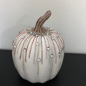 May include: A white decorative pumpkin with a brown stem, adorned with silver and red beaded lines and sparkling clear crystals. This fall-themed accent piece is perfect for seasonal home decor.