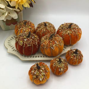 May include: A collection of decorative pumpkins in various sizes, perfect for fall. The pumpkins are covered in orange and red velvet and adorned with gold and silver beads. Each pumpkin has a small, dark brown stem.